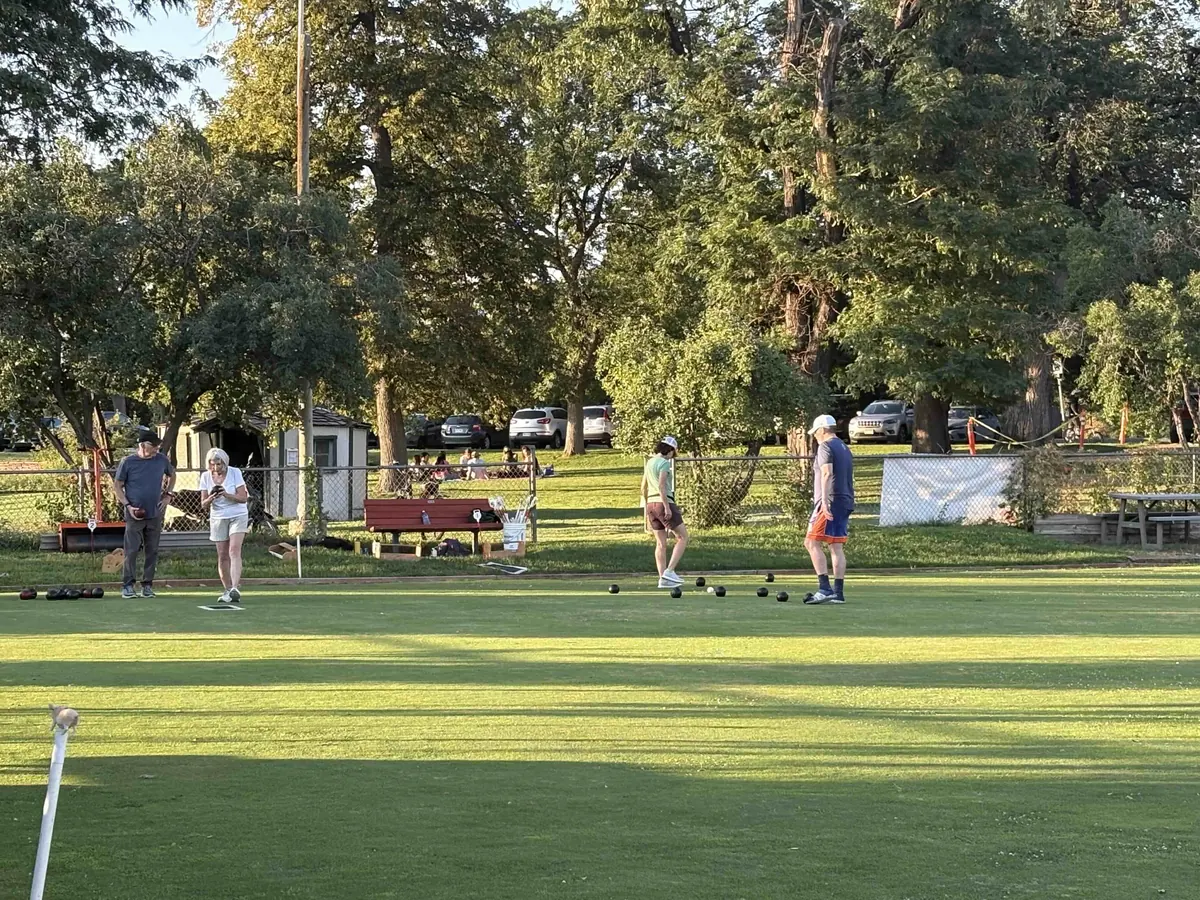 A summer evening on the Washington Park green: four bowlers in shorts and caps deliver bowls toward the jack beneath tall shade trees, with red benches and the clubhouse shed behind
