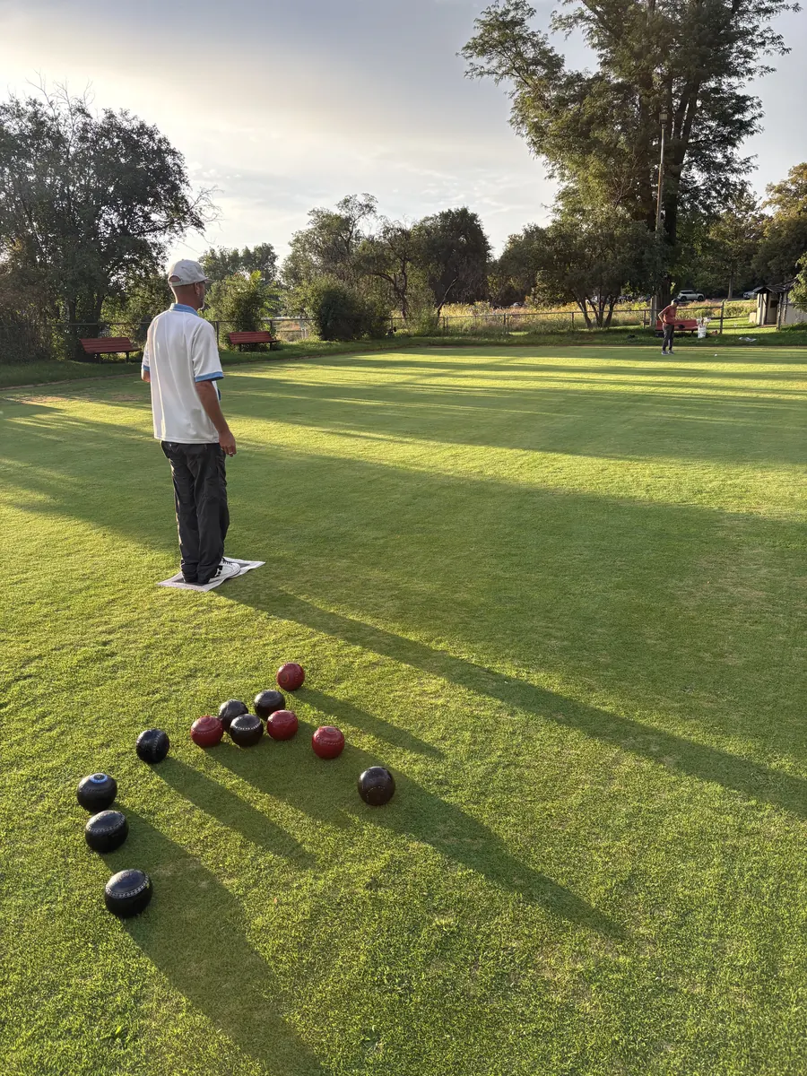 Late-afternoon sun casts long shadows across the green as a bowler in a white cap stands on the mat beside a cluster of black and red bowls