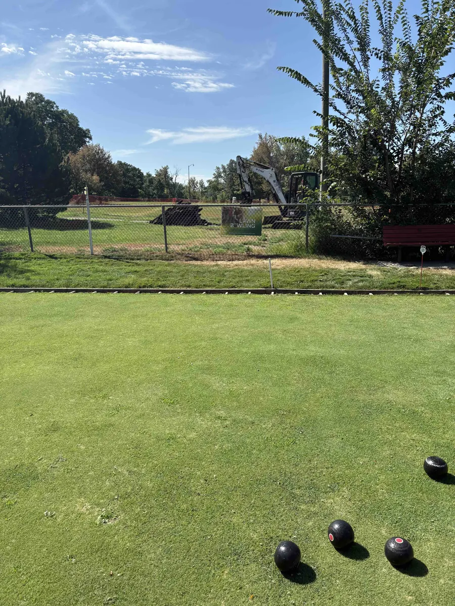 Four black bowls rest on the closely mown green, with a chain-link fence, a red bench and the mature trees of Washington Park beyond on a clear blue day